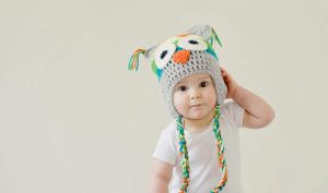 A cute baby wearing an owl hat standing in a basket, smiling and playful.